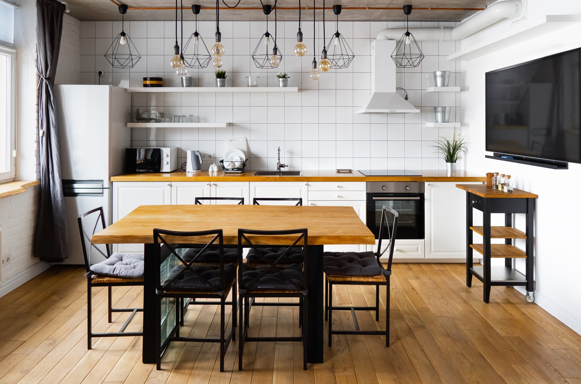 Moderno escandinavo un diseño interior de cocina comedor con gran mesa de madera y sillas contra suelo de madera clara, paredes blancas brillantes y muebles con TV, electrodomésticos y bombillas colgantes